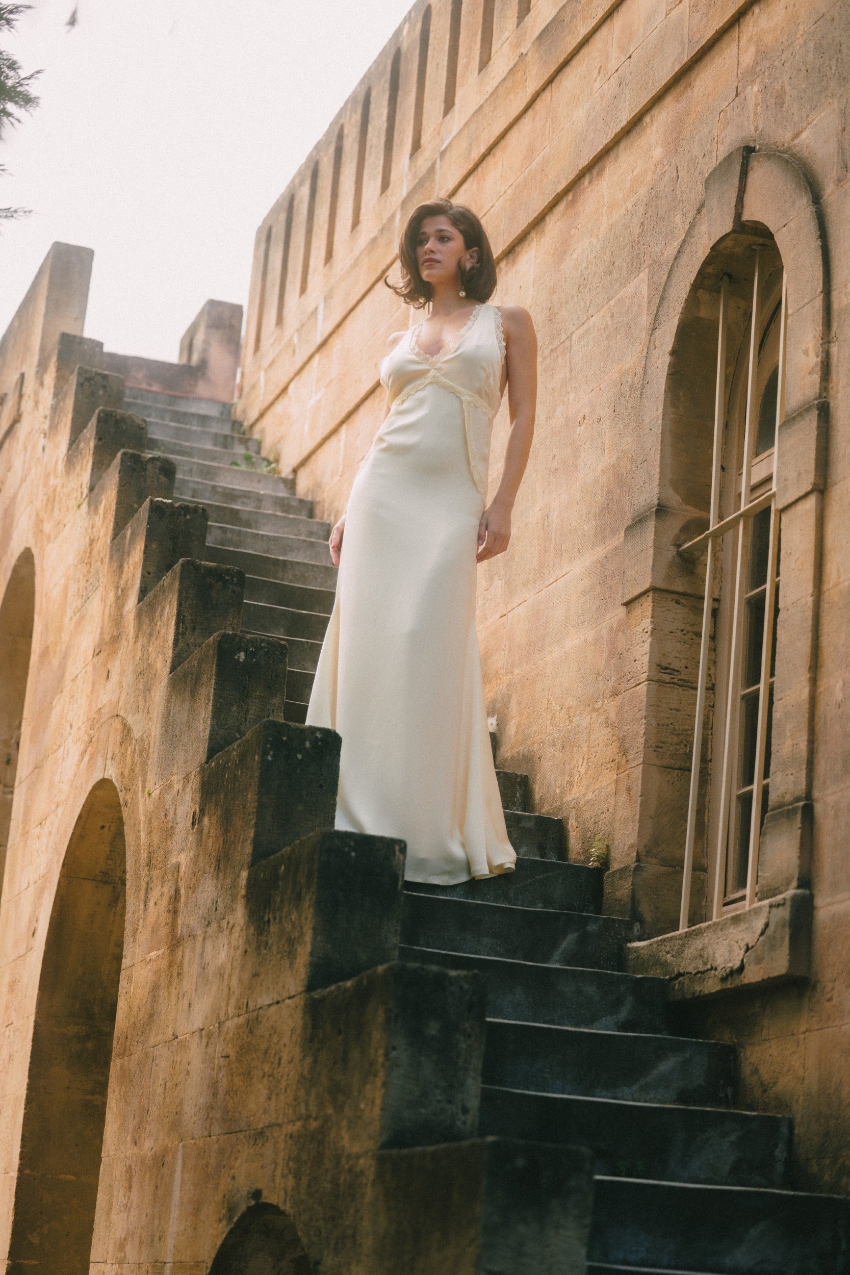 A woman wearing a sleeveless white wedding gown stands on dark stone stairs beside a tan stone building with arched windows.